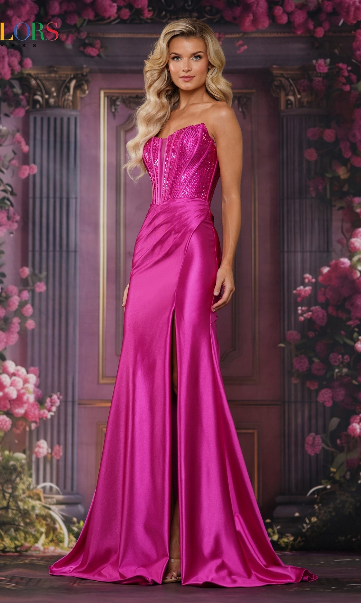 A woman models the Lace-Up Long Corset Prom Dress: Colors Dress 3692, featuring a hot pink satin skirt with a thigh-high slit and a sparkling beaded corset bodice, set against a floral and column backdrop.